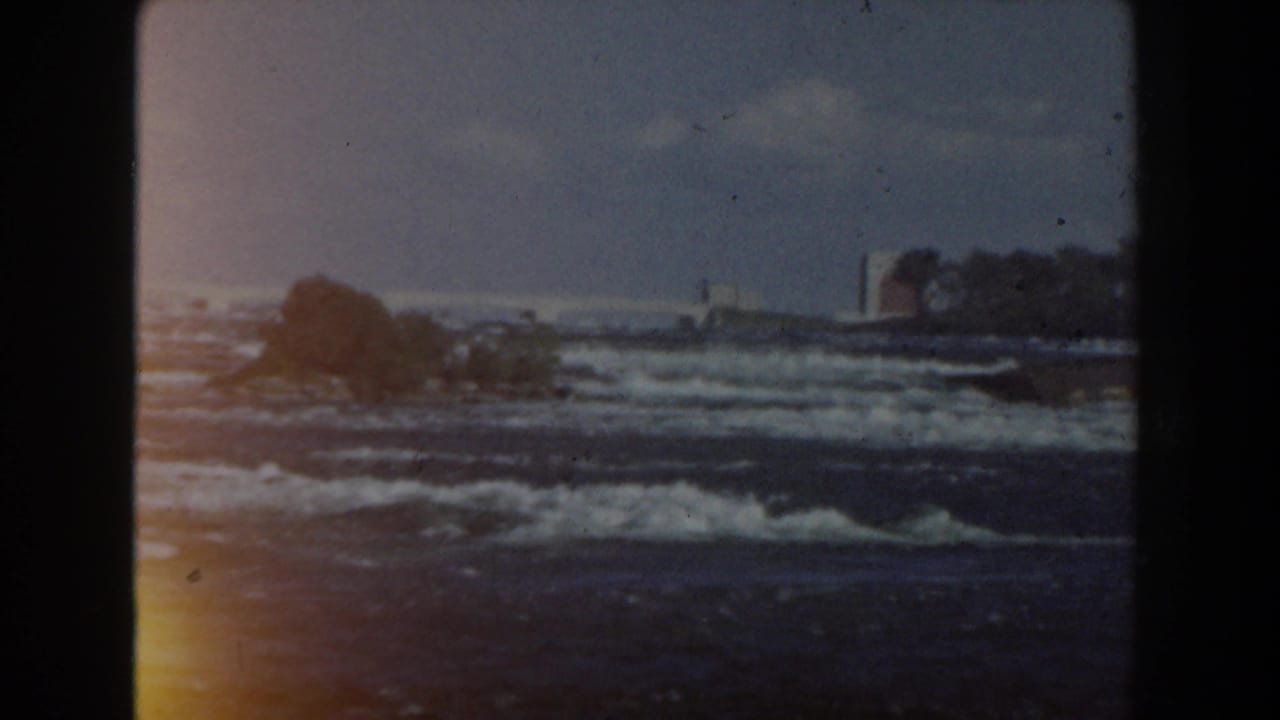 Waves roll toward a dark shoreline with trees and pale sky, framed by 8mm film borders.