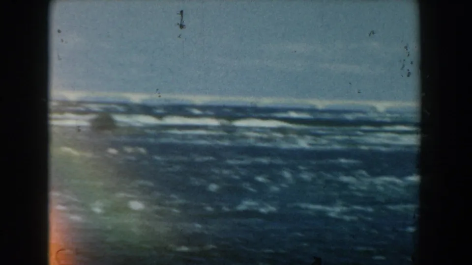 1959 Michigan 8mm frame showing choppy blue shoreline waves beneath a pale sky, with film grain, dust, and gate edges visible.