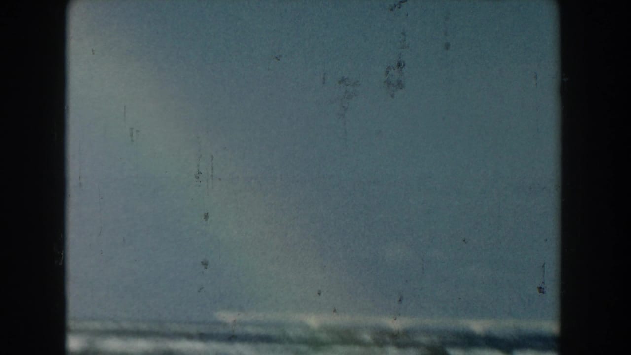 1959 Michigan 8mm frame with a low horizon of waves beneath open blue sky, marked by light scratches and home-movie grain.