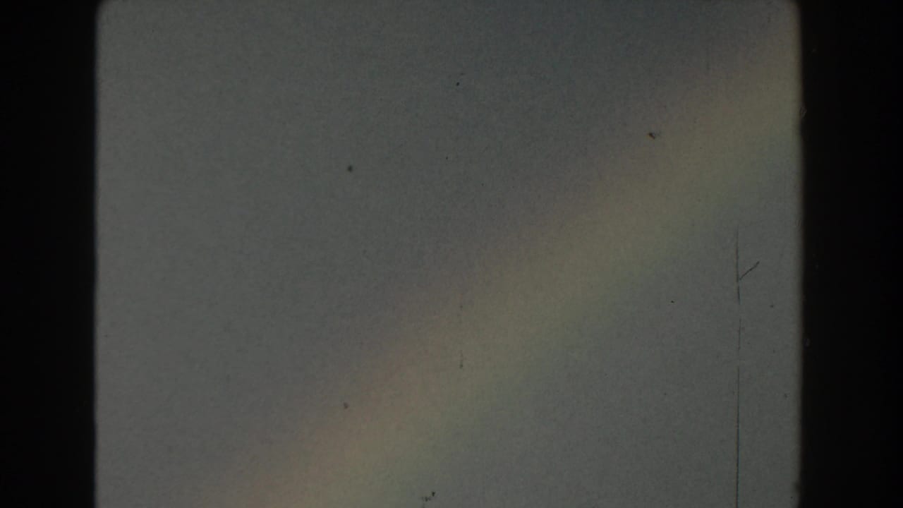 Faint rainbow in a cloudy Michigan sky from 1959 8mm home-movie footage, framed by dark gate edges and visible film wear.