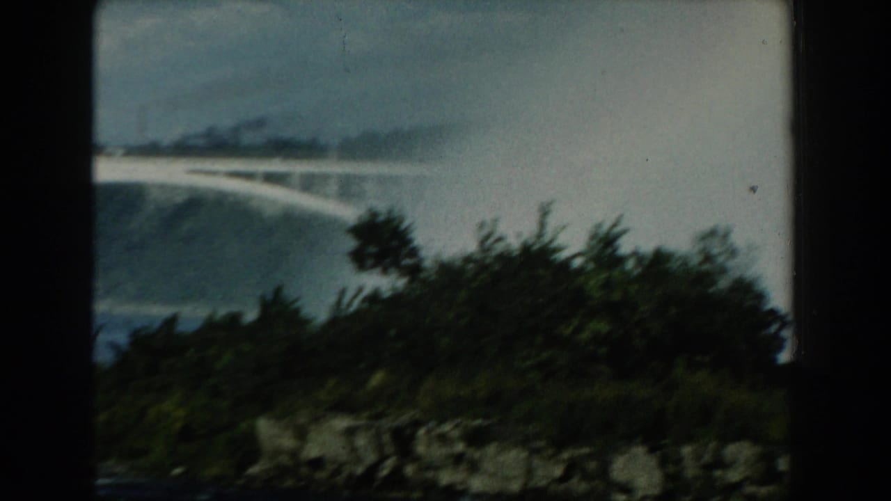 Faint rainbow over water with trees and rocky shoreline in the foreground.