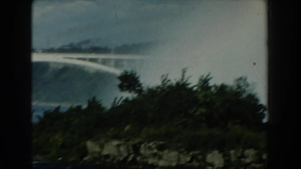 1959 Michigan 8mm frame showing a faint rainbow above water, partly obscured by dark trees and a rocky shoreline.