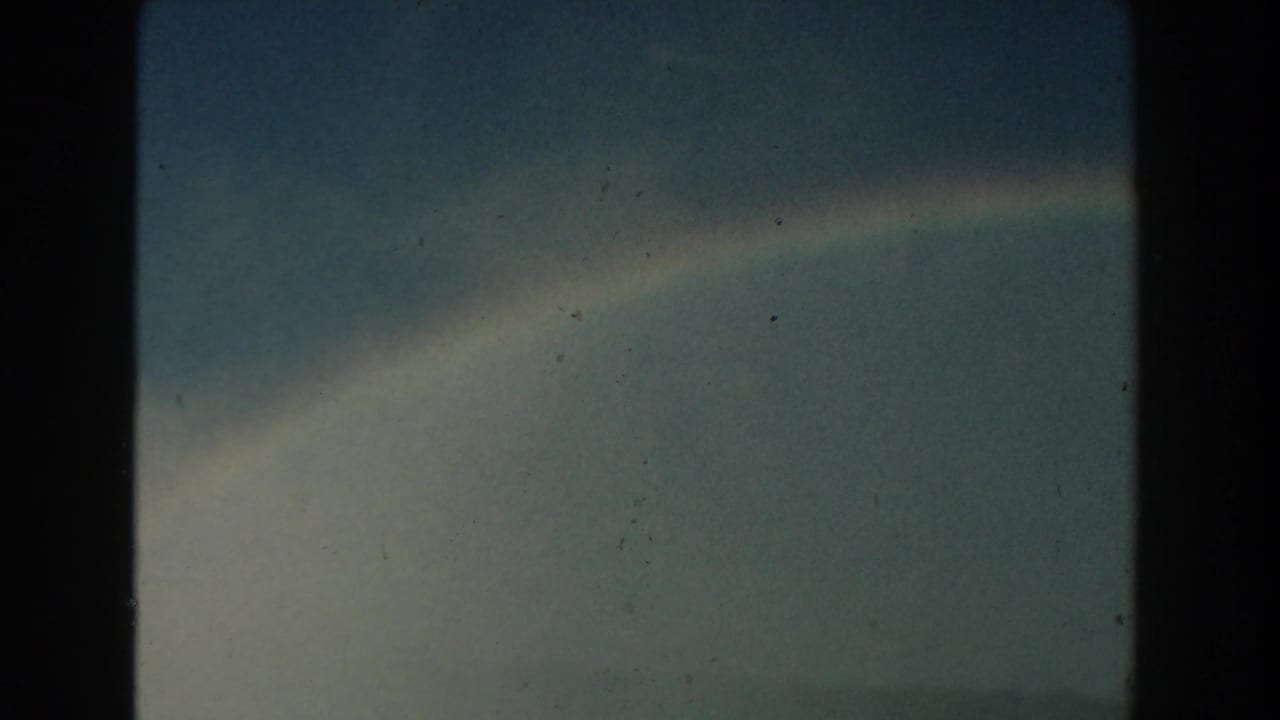 1959 Michigan 8mm frame showing a faint rainbow curving through a muted blue-gray sky with visible film grain.