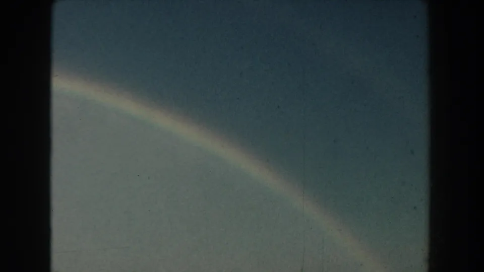 1959 Michigan 8mm frame of a rainbow arc fading through a cloudy blue-gray sky, with dust and grain from the film scan.