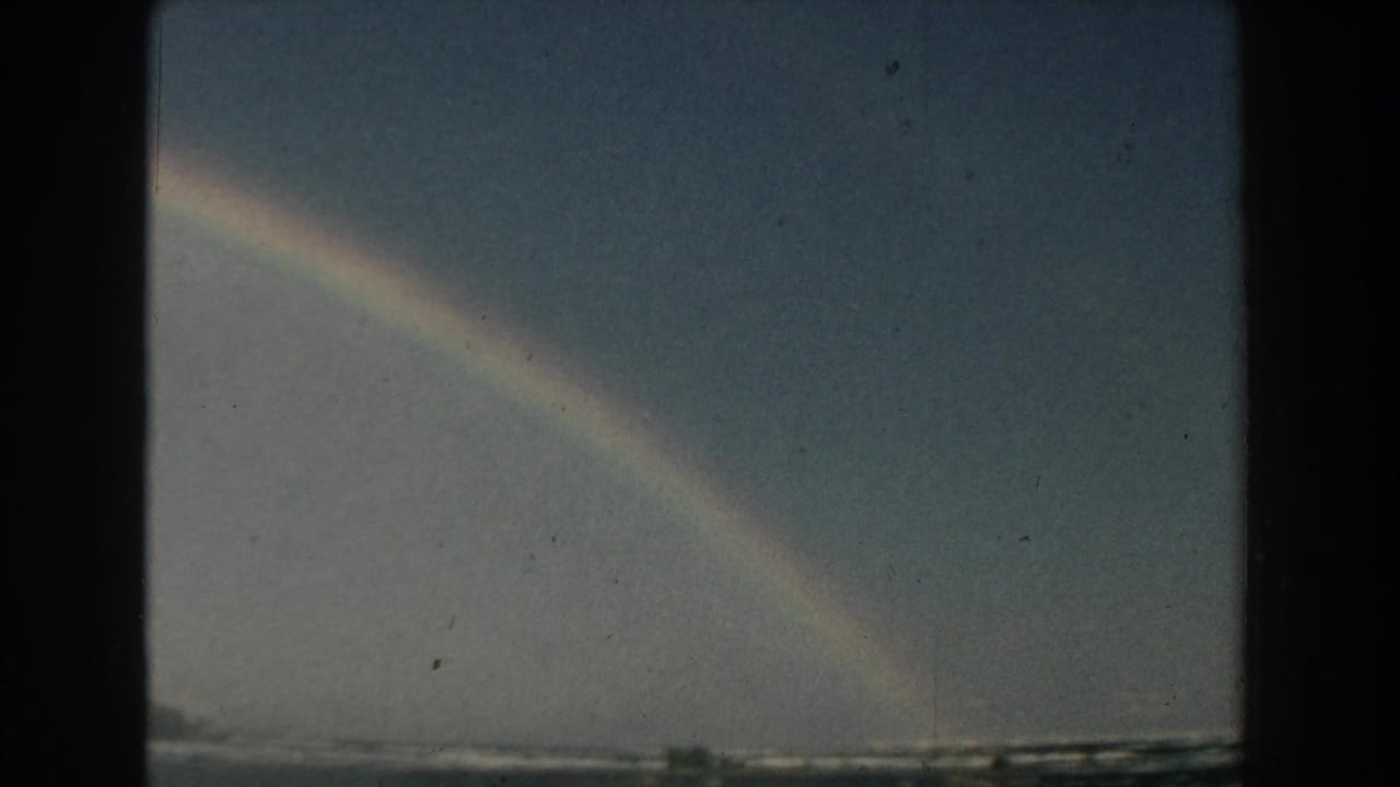 Rainbow over choppy water with a low distant shoreline under blue sky.