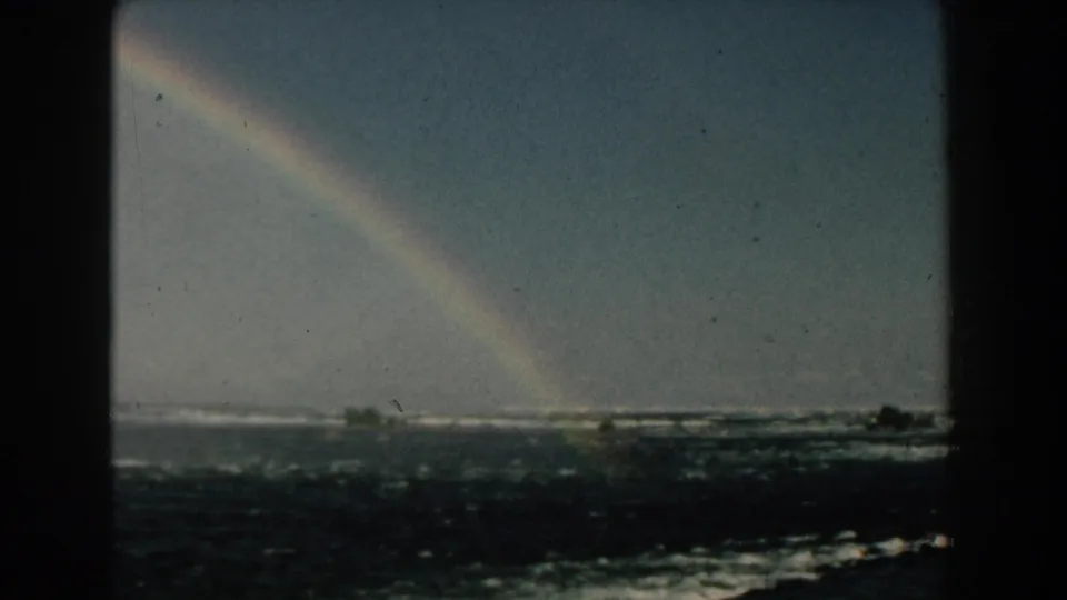 1959 Michigan 8mm frame with a bright rainbow above foamy waves and a low, tree-lined horizon.