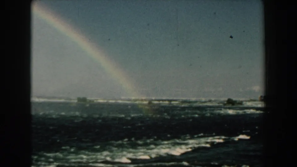1959 Michigan home-movie frame showing a rainbow above rough, foaming water and a low shoreline in the distance.