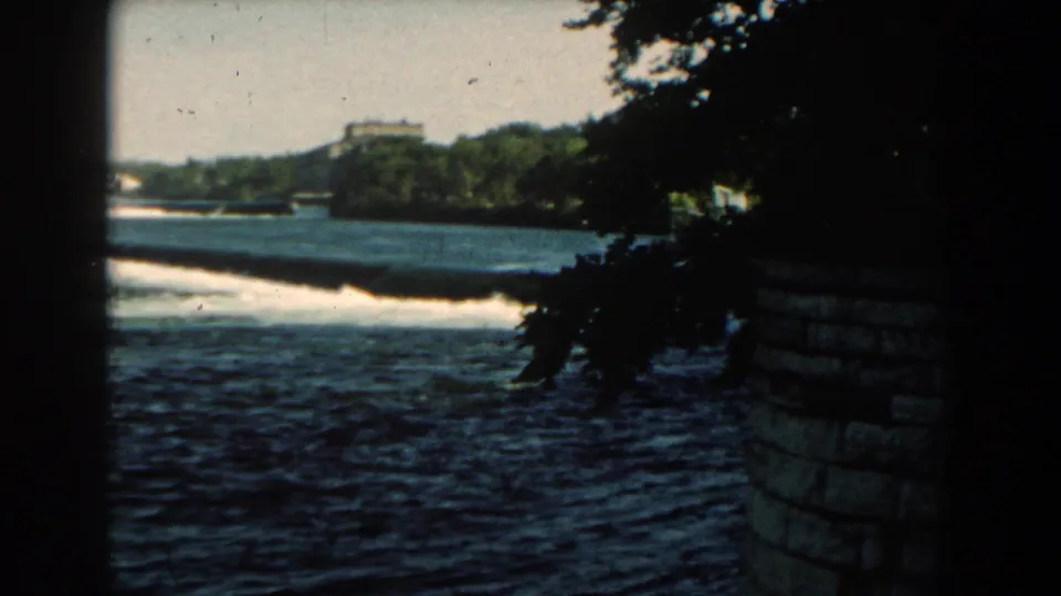 1959 Michigan 8mm frame showing choppy shoreline water, trees, a distant structure, and a stone wall or embankment at right.