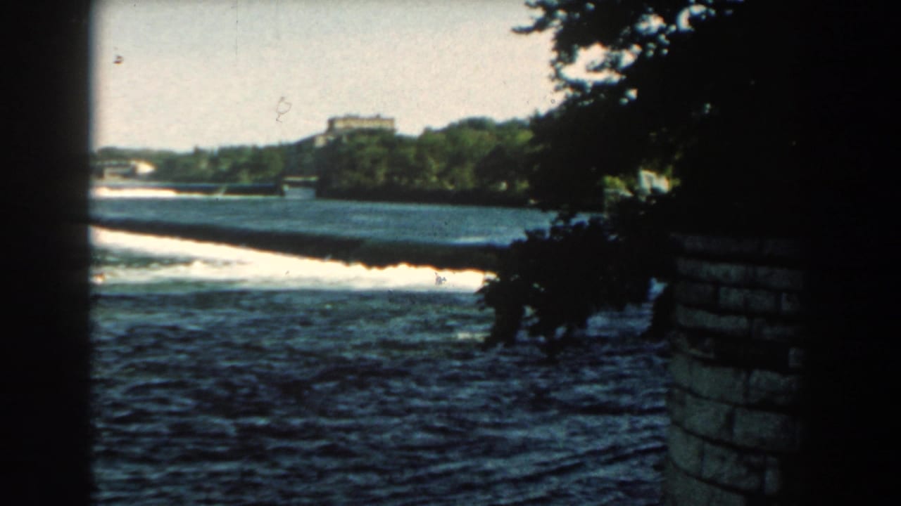 1959 Michigan 8mm view of a river spilling over a long low dam, framed by shadowed foliage and a structural edge.