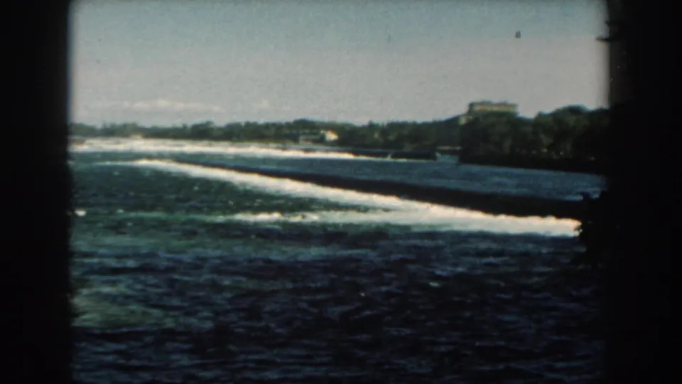 A wider 1959 Michigan home-movie frame shows the dam stretching across the river with rapids and a distant structure on the far shore.