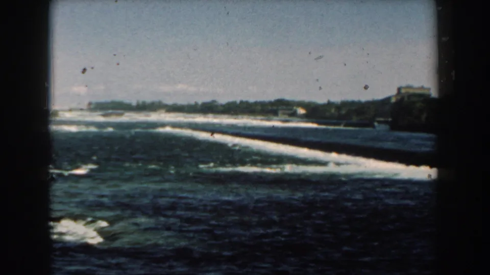 Late-frame 8mm view of the Michigan river dam, with dark foreground shade, churning water, trees, and distant shoreline forms.