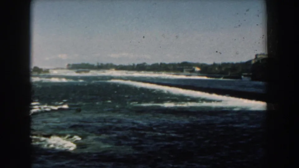 Brief 1959 Michigan 8mm view of a shoreline with breaking waves, distant structures, and heavy dark frame borders.