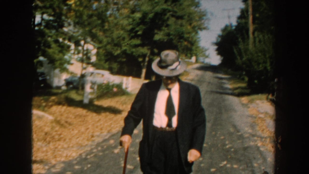 Man in dark suit and grey hat walks down a sunny residential street with a cane.