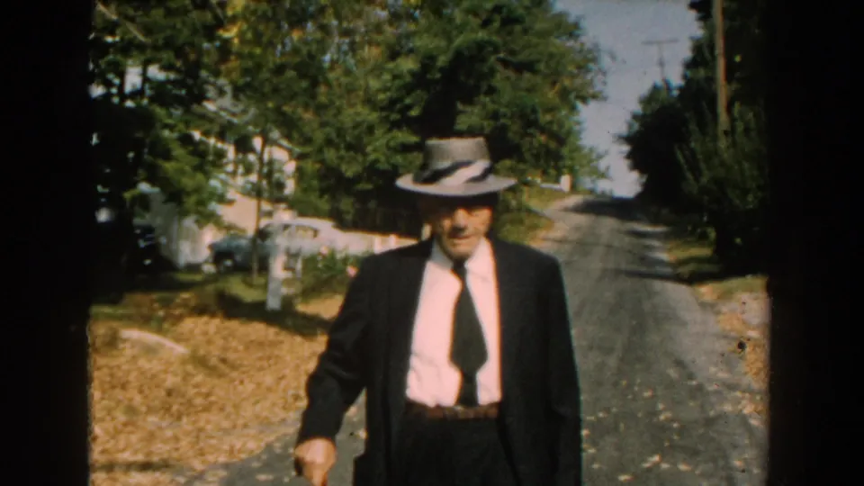 The man in a dark suit and grey fedora approaches closer on the Michigan residential street, holding a brown cane beside dry grass and trees.