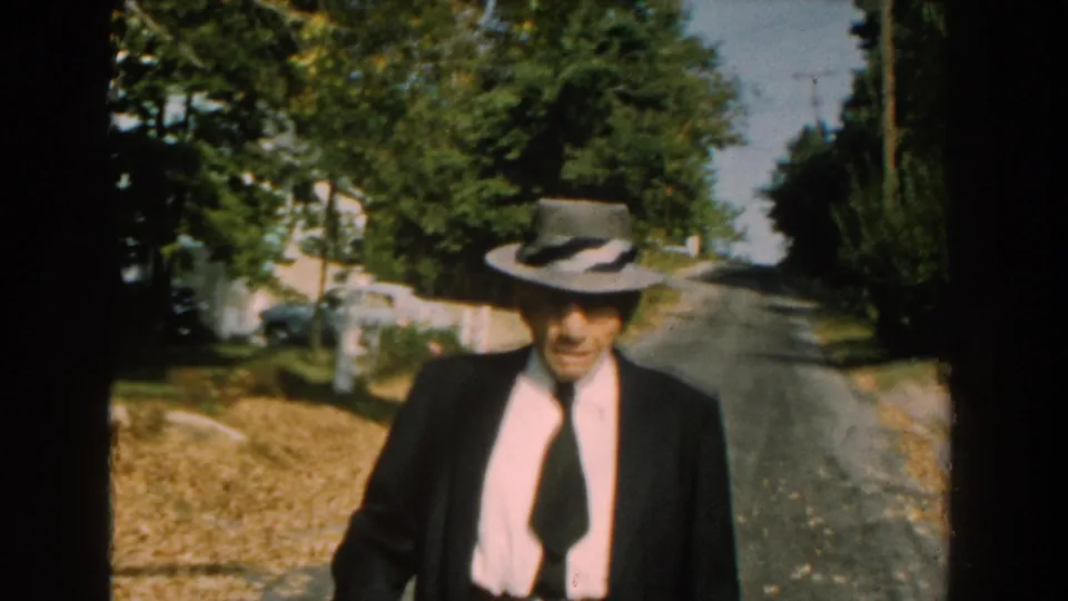 A closer 1959 Michigan frame shows the suited man in a grey hat walking toward the camera with trees, road, and residential fencing behind him.