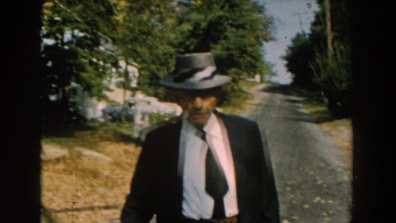 In 1959 Michigan home-movie footage, an older man in a fedora, dark suit, white shirt, and tie walks toward the camera on a tree-lined road.