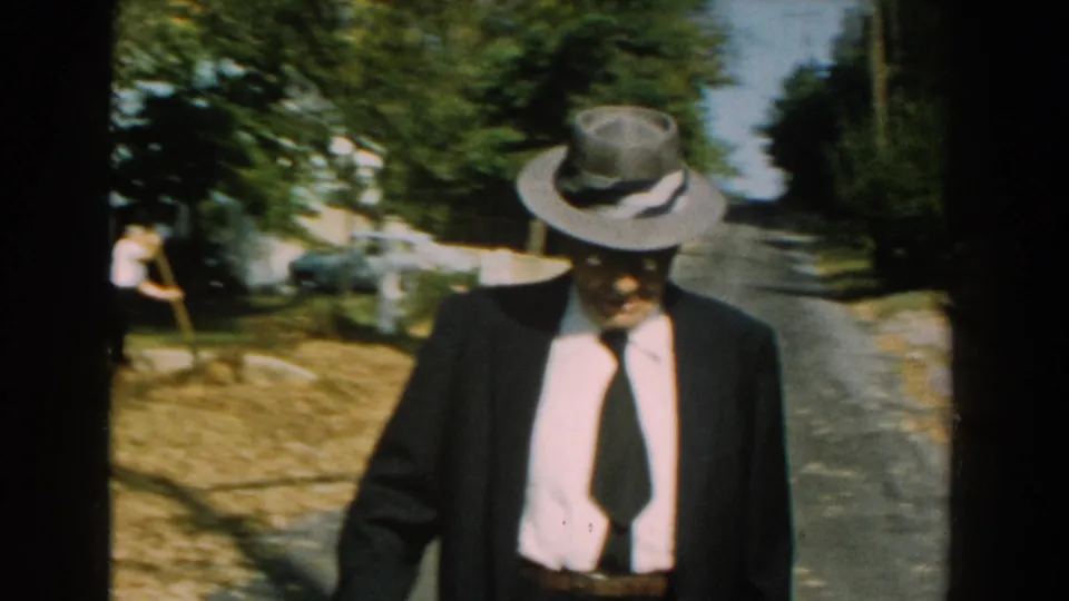 The older man draws closer to the lens, his fedora and suit prominent against the sunlit road and leafy residential background.