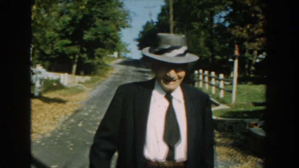 The man appears to smile while walking past the camera, with a white fence, trees, and the residential road visible behind him.