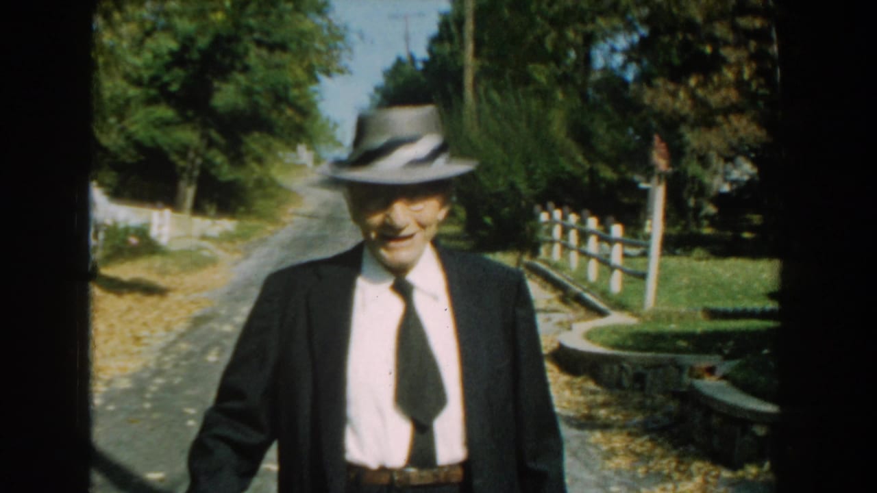 Elderly man in a fedora and dark suit walks along a rural road toward the camera.