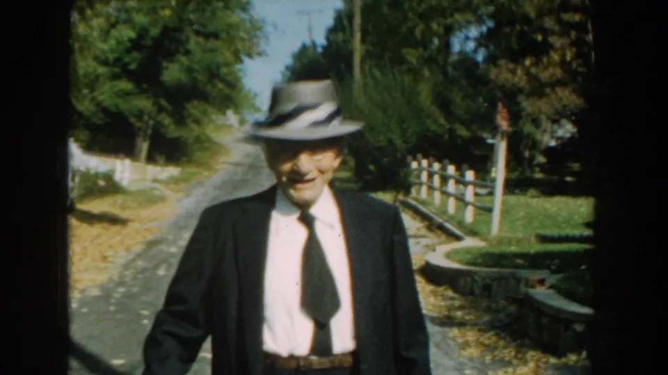 In 1959 Michigan home-movie footage, an elderly man in a suit and fedora approaches along a tree-lined rural road.