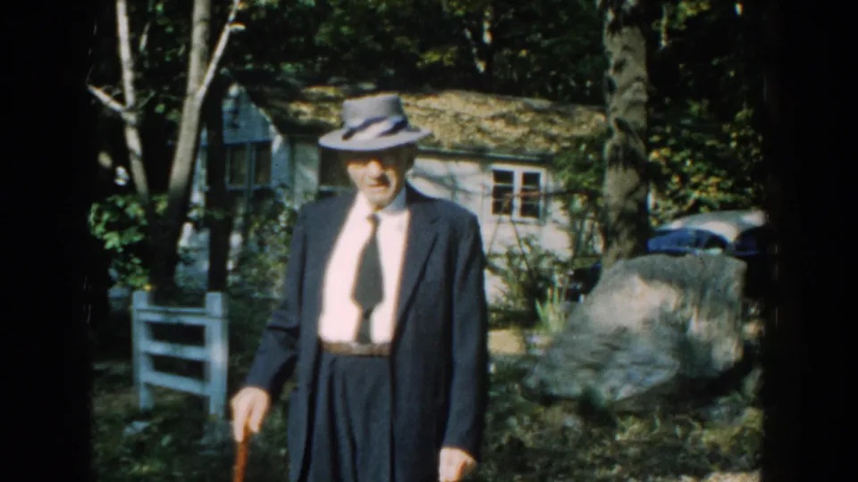 An elderly man in a dark suit and fedora carries a cane in a wooded Michigan yard near a small cottage.