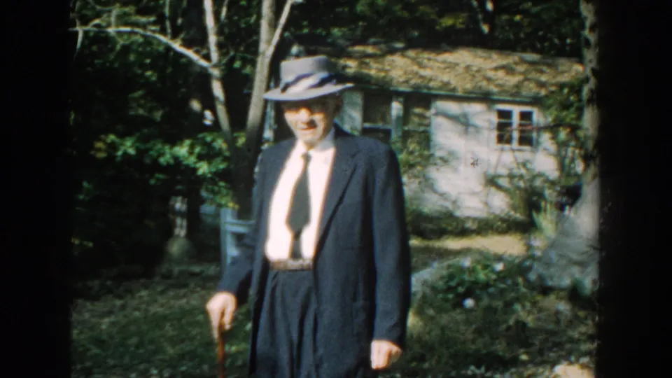 The suited elderly man moves through a wooded yard with a cane, with a white fence and cottage behind him.
