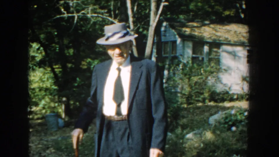 A smiling elderly man in 1959 Michigan footage faces the camera while holding a cane in a shaded cottage yard.