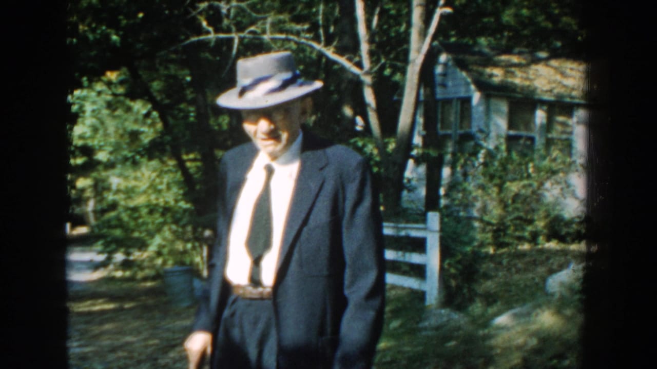 In 1959 Michigan home-movie footage, an elderly man in a dark suit, tie, and fedora walks with a cane near trees and a house.