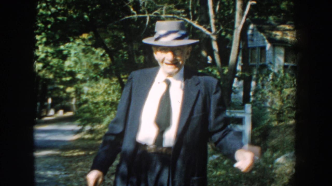 The elderly man smiles toward the camera in this 1959 Michigan 8mm frame, walking with a cane near a fence and house exterior.