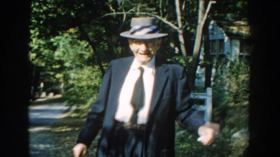 The elderly man smiles toward the camera in this 1959 Michigan 8mm frame, walking with a cane near a fence and house exterior.