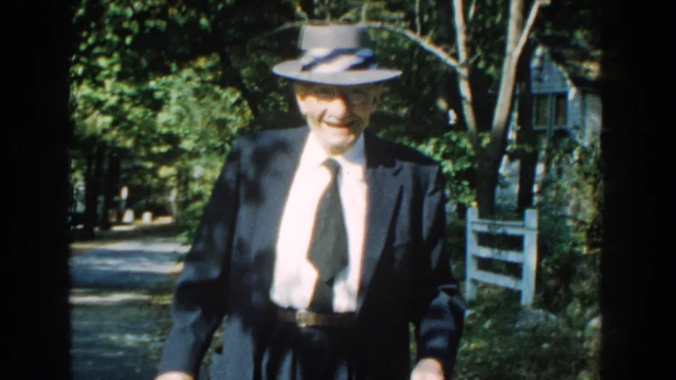 A closer 1959 Michigan home-movie frame shows the elderly man in formal clothes and fedora near trees, a path, and a rustic house.