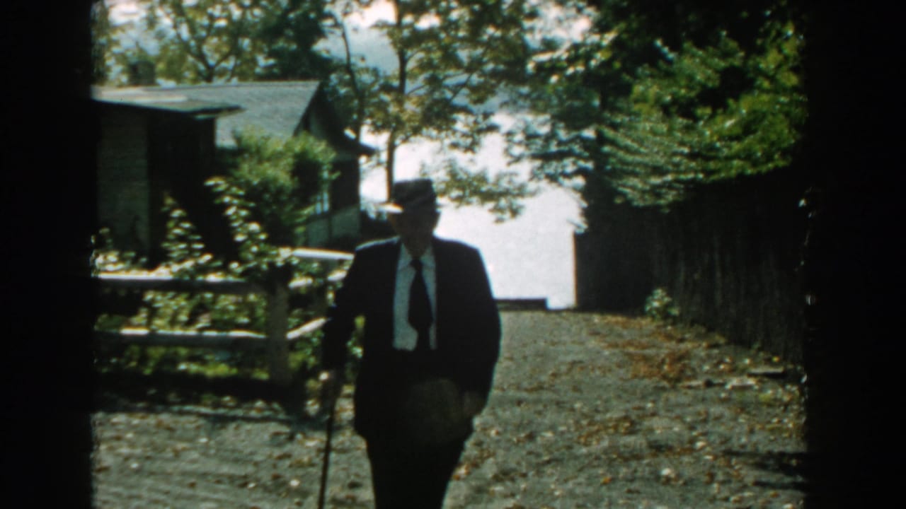 In 1959 Michigan home-movie footage, an elderly man in a dark suit, tie, and fedora walks toward the camera with a cane.