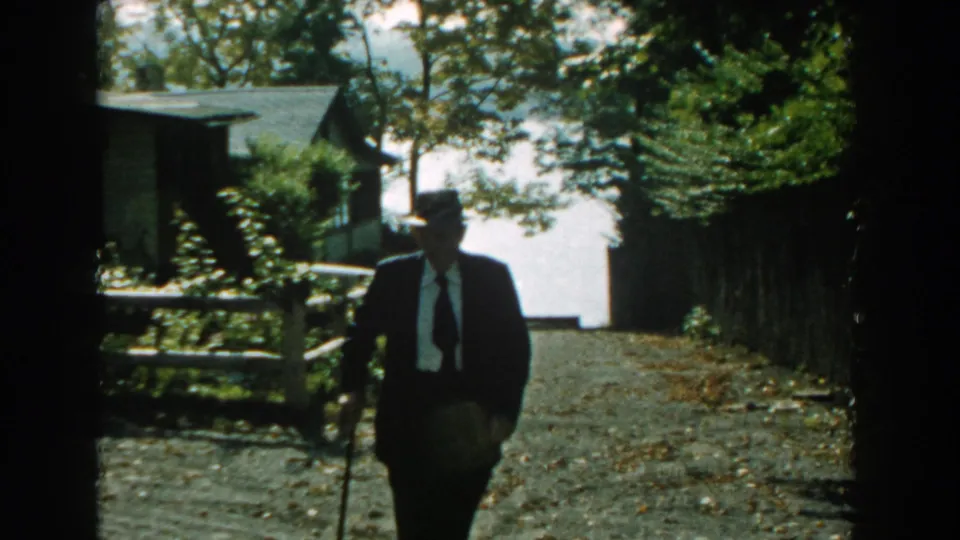 In 1959 Michigan home-movie footage, an elderly man in a dark suit, tie, and fedora walks toward the camera with a cane.