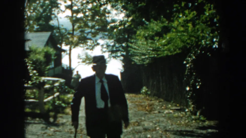 The elderly man continues toward the camera on a tree-lined gravel path, with a fence and house visible behind him.