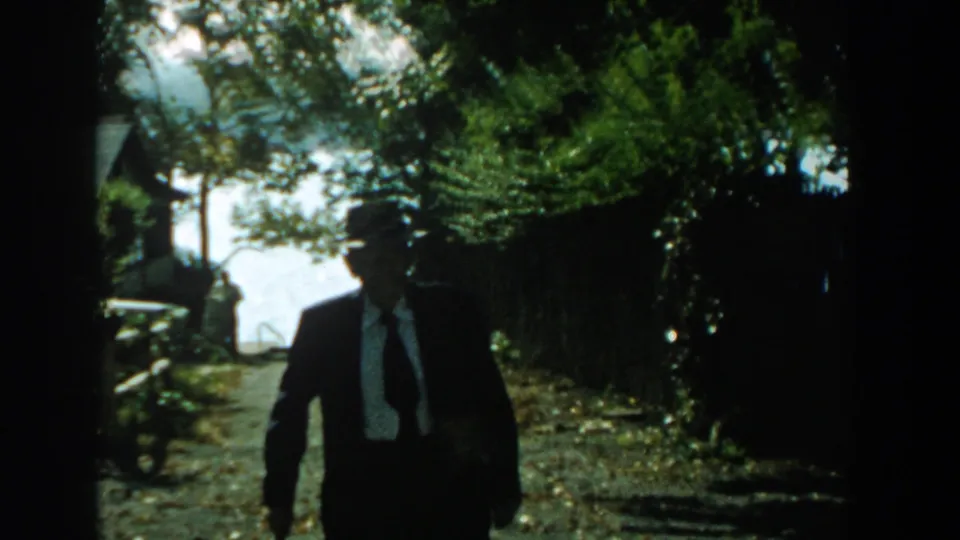 The man in a dark suit and fedora is closer to the camera, partly shaded by foliage as he walks with his cane.