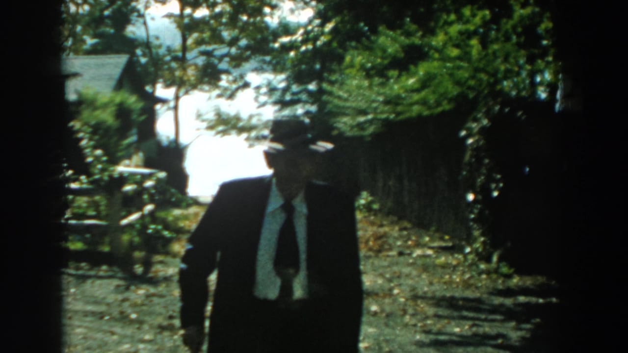 In 1959 Michigan home-movie footage, a man in a dark suit and grey fedora walks toward the camera on a bright outdoor path.