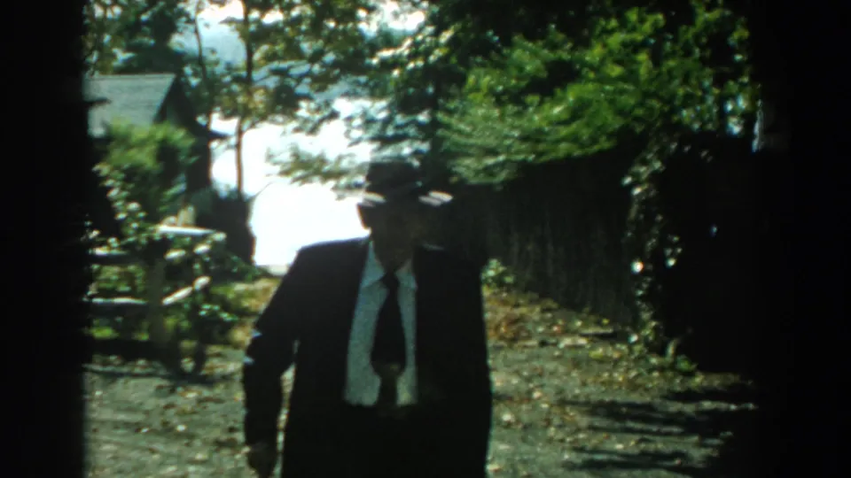 In 1959 Michigan home-movie footage, a man in a dark suit and grey fedora walks toward the camera on a bright outdoor path.