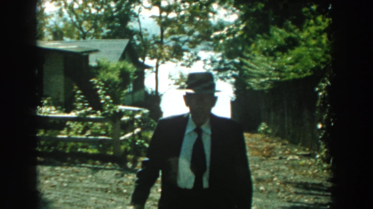Suited man in a fedora closer to the camera, with bright trees and a small building behind him.