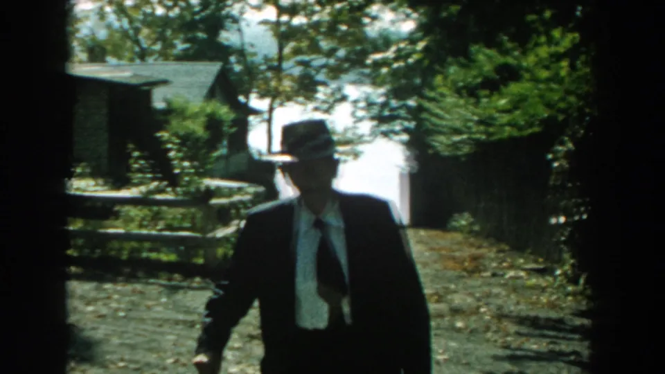 A formally dressed man nears the camera on a sunny path in 1959 Michigan home-movie footage, with fence and house visible behind.