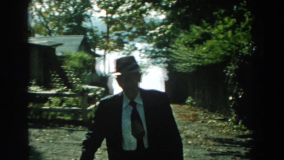 Near the end of the clip, the man in a dark suit and fedora approaches the camera, backlit by a sunny Michigan yard in 1959.