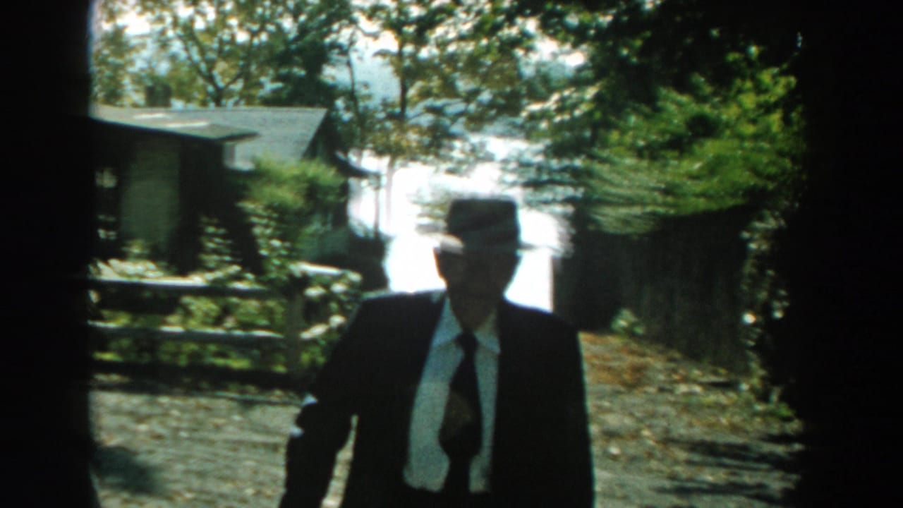 Older man in a dark suit and fedora walks along a gravel path in a leafy yard.