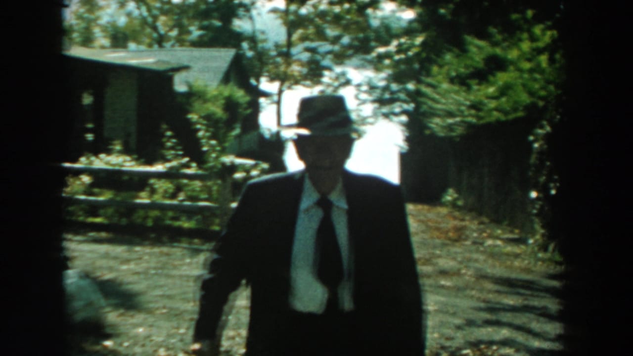 Older man in suit and fedora approaches the camera near trees and a fence.
