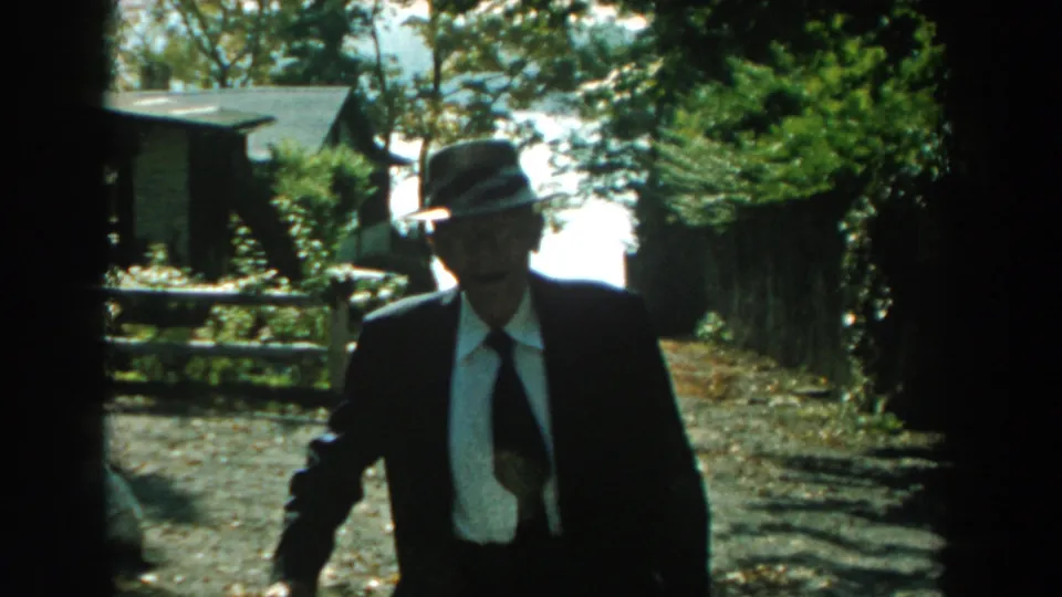 The man is closer to the camera, his suit, white shirt, tie, and fedora visible against the shaded path and suburban yard.