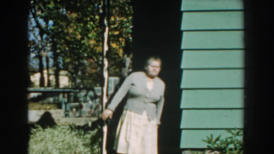 A woman in a light cardigan and skirt stands near the side of a house in the same Michigan home-movie sequence.