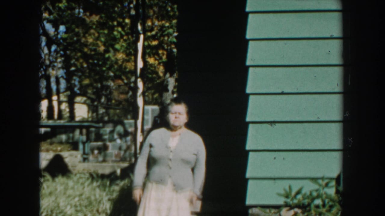 Older woman standing near a porch beside green house siding.