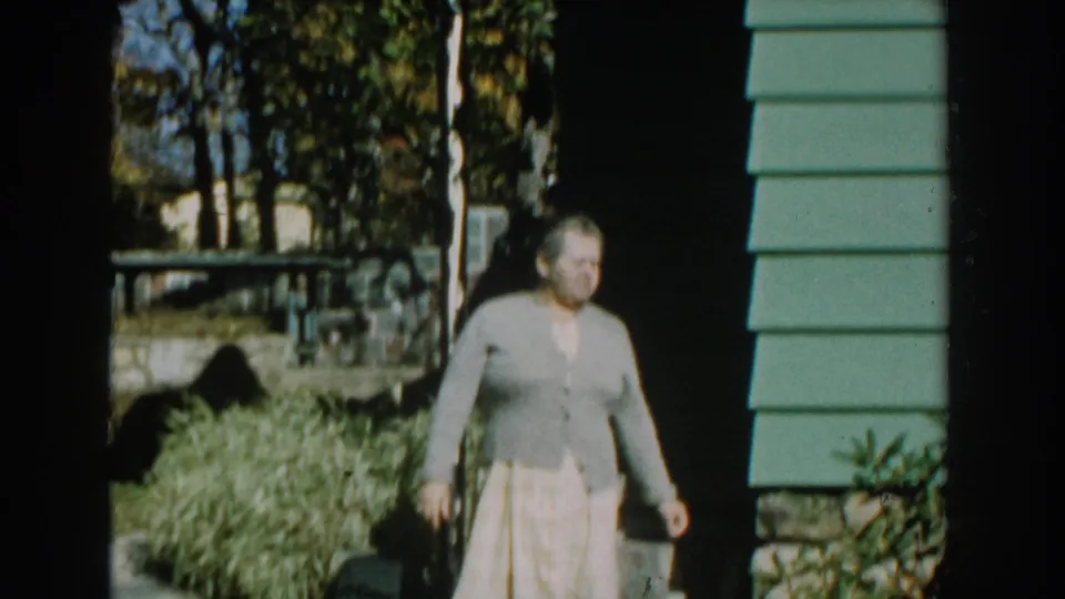 The woman steps out from the shaded porch area of a green-sided Michigan house, with shrubs and trees behind her.
