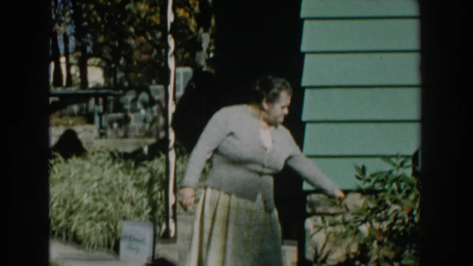The woman bends slightly and extends her left hand toward a shrub near the steps, with green siding and yard foliage visible.