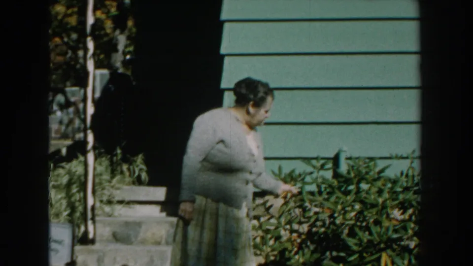 Near the green-sided house, the woman leans over a garden shrub as if picking or inspecting something in the plants.