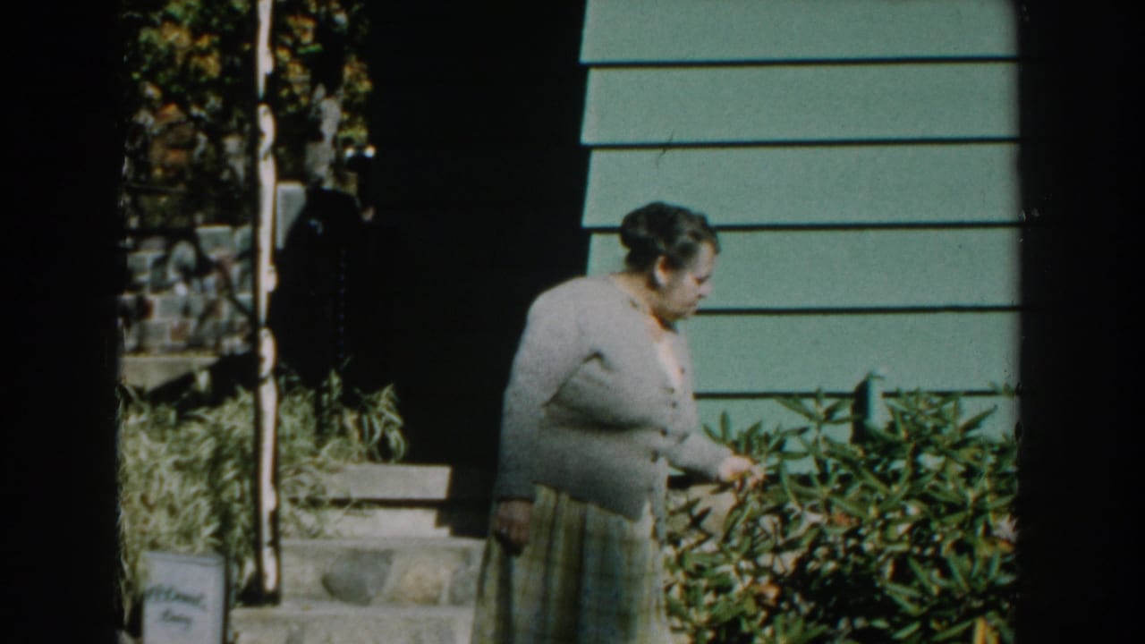 Woman in cardigan reaches toward shrubs beside green house siding and porch steps.