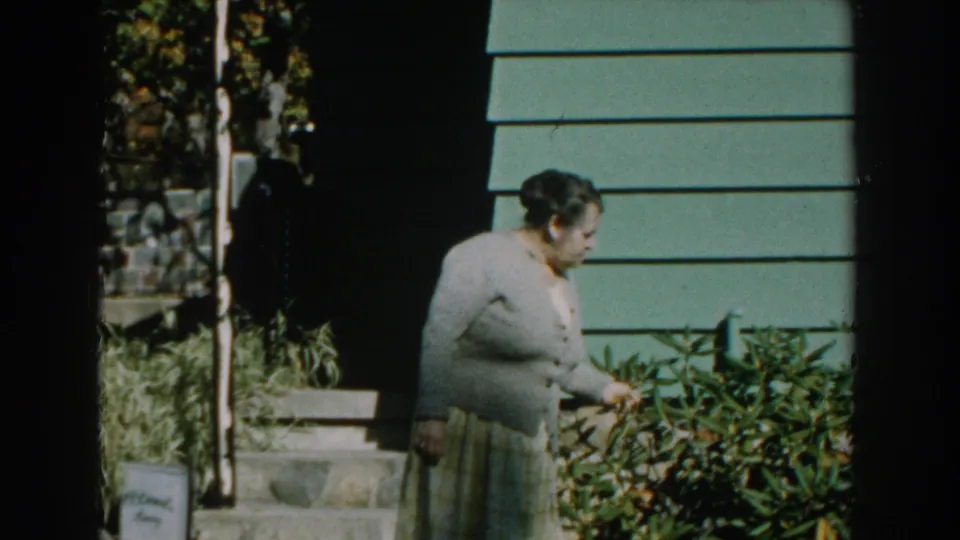 In 1959 Michigan home-movie footage, a woman in a light cardigan and plaid skirt tends shrubs outside a green-sided house.